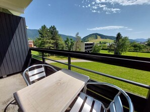 A balcony with a table and chairs, offering a view of a green landscape with trees and hills.