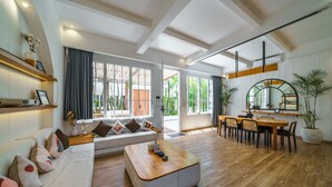 Uma sala de estar moderna com sofá, mesa de centro, mesa de jantar e cadeiras. Há grandes janelas com cortinas, um vaso de planta e um espelho na parede.