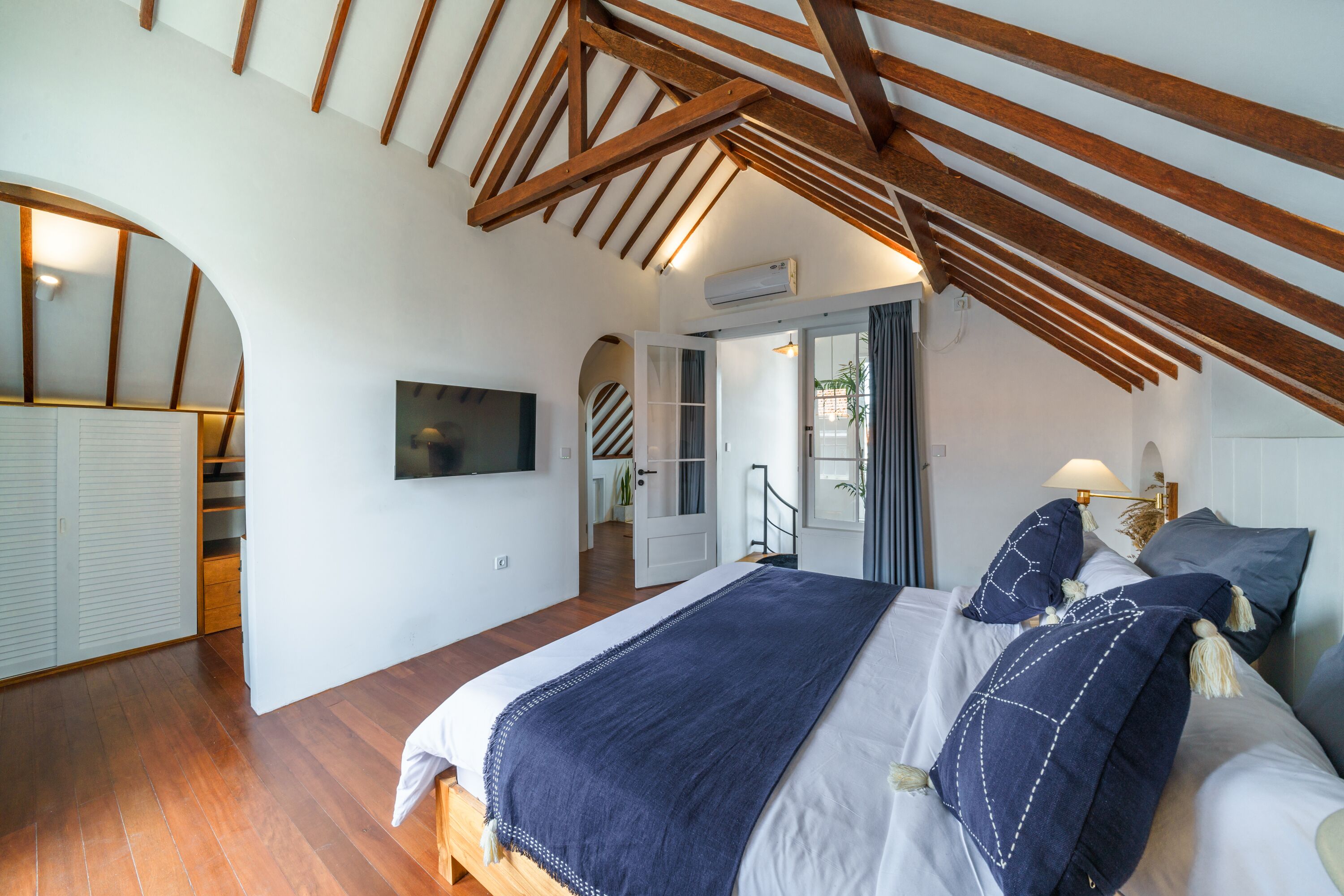 Ein Schlafzimmer mit Holzbalkendecke, einem Bett mit blauer Decke, einem Fernseher an der Wand und einer Treppe, die zu einem Balkon führt.