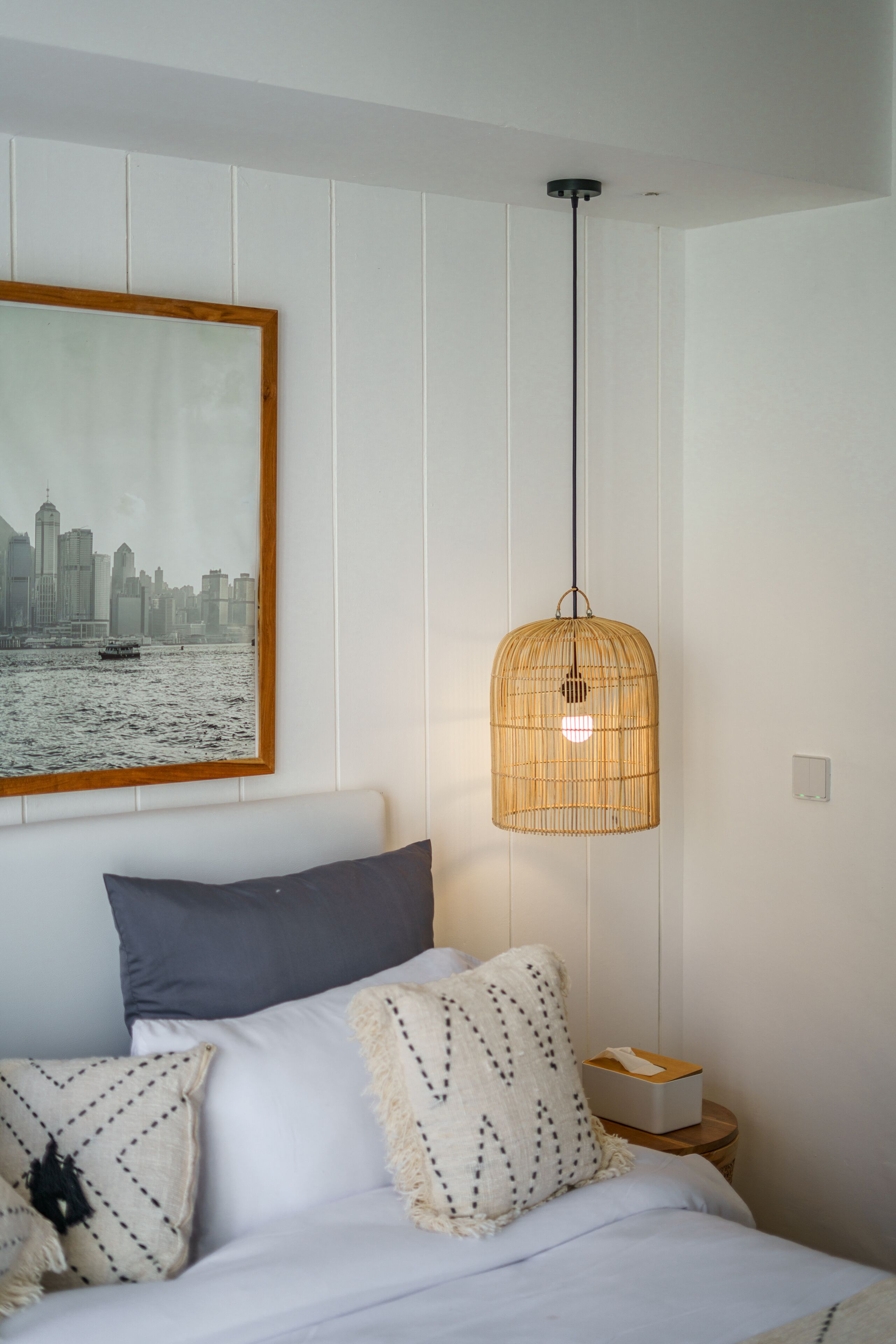 A neatly arranged bedroom with a bed, pillows, a bedside table with a tissue box, a hanging light fixture, and a cityscape painting on the wall.