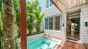 Uma área de piscina com deck de madeira, rodeada por vegetação exuberante e um edifício branco com grandes janelas.