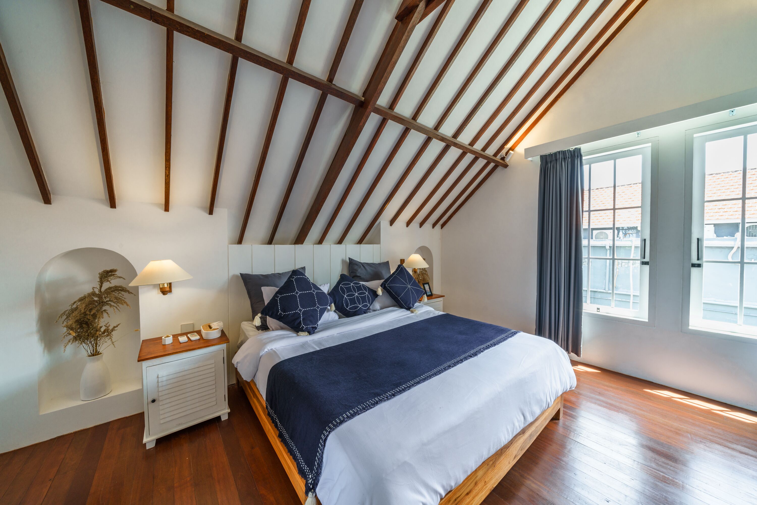 A bedroom with a sloped ceiling, a large bed with a navy blue bedspread, wooden headboard, bedside tables with lamps, and a window with curtains.