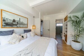 Un dormitorio moderno con una cama grande, un cuadro de un paisaje urbano, una planta en maceta y vistas a un baño.