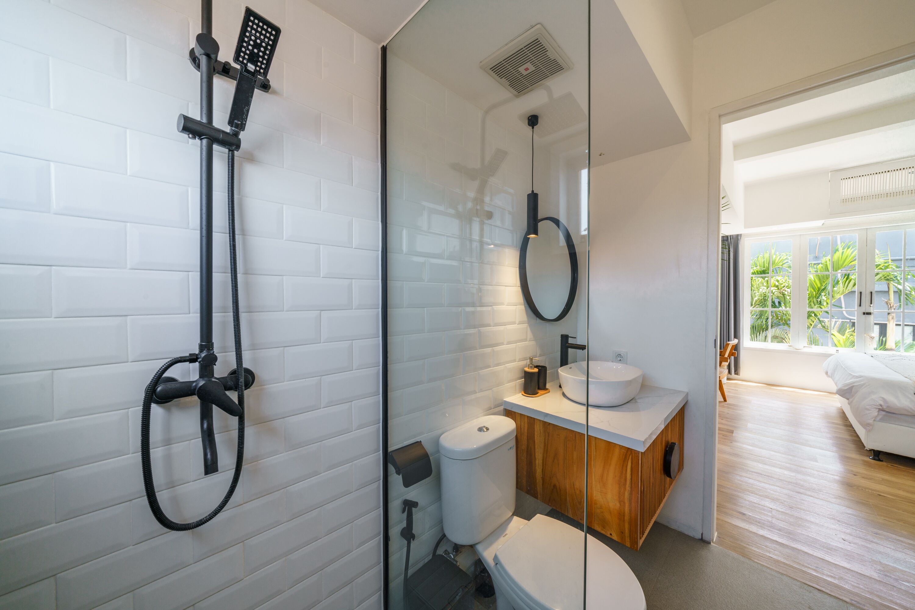 A modern bathroom with a glass shower enclosure, white tiled walls, a wooden vanity, and a view of a balcony with plants.