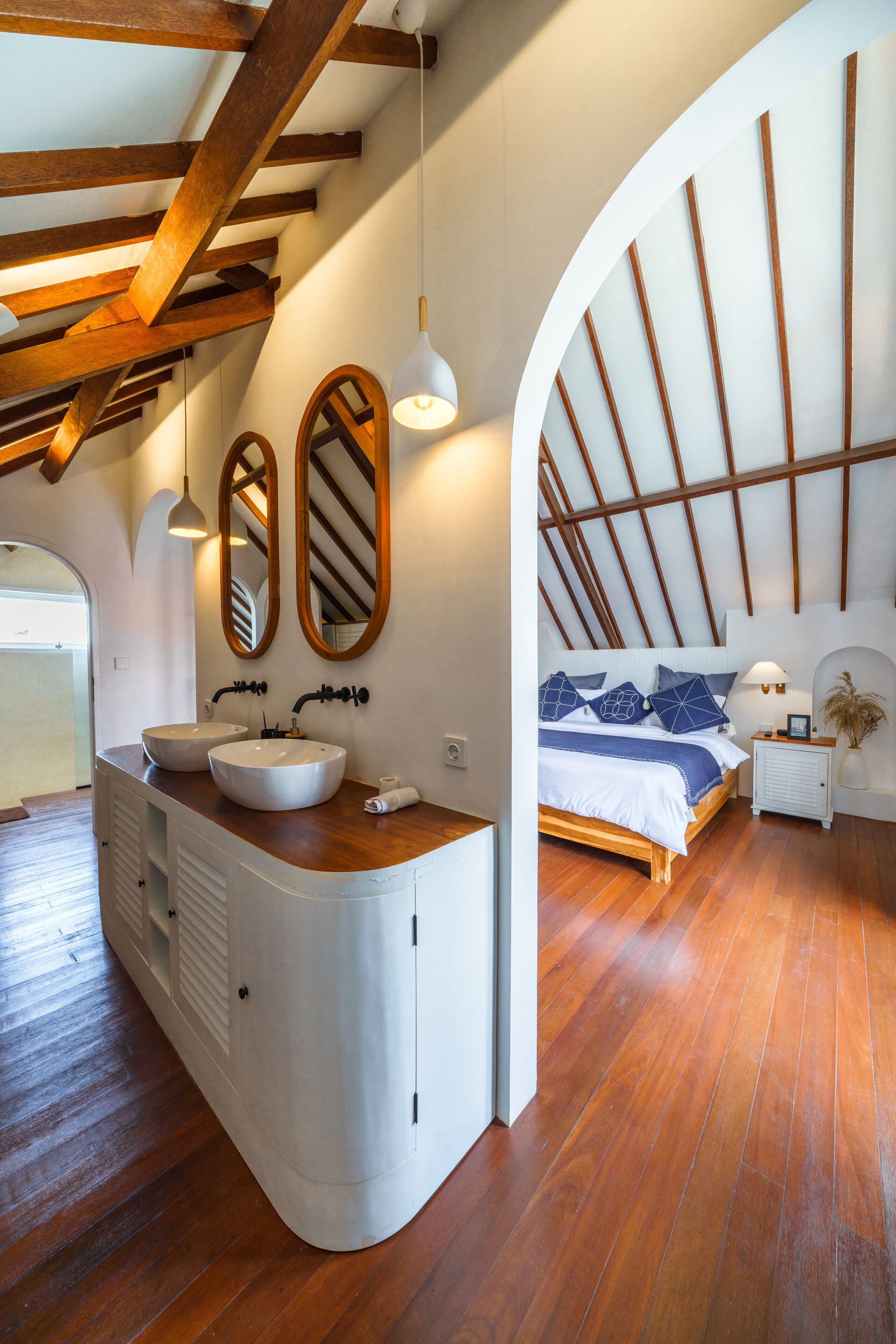 A bedroom with a curved vanity, two mirrors, a bed with blue bedding, and wooden beams on the ceiling.