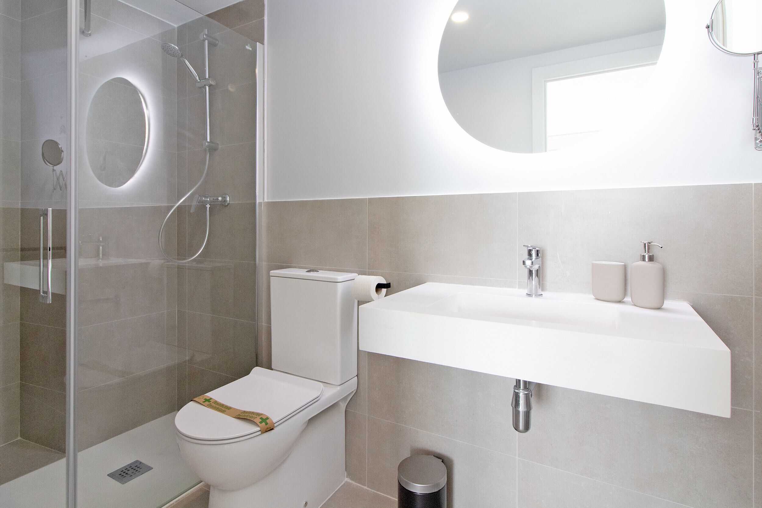 Un baño moderno con ducha envuelta en vidrio, inodoro con dispensador de toallas de papel y lavamanos con espejo superior.