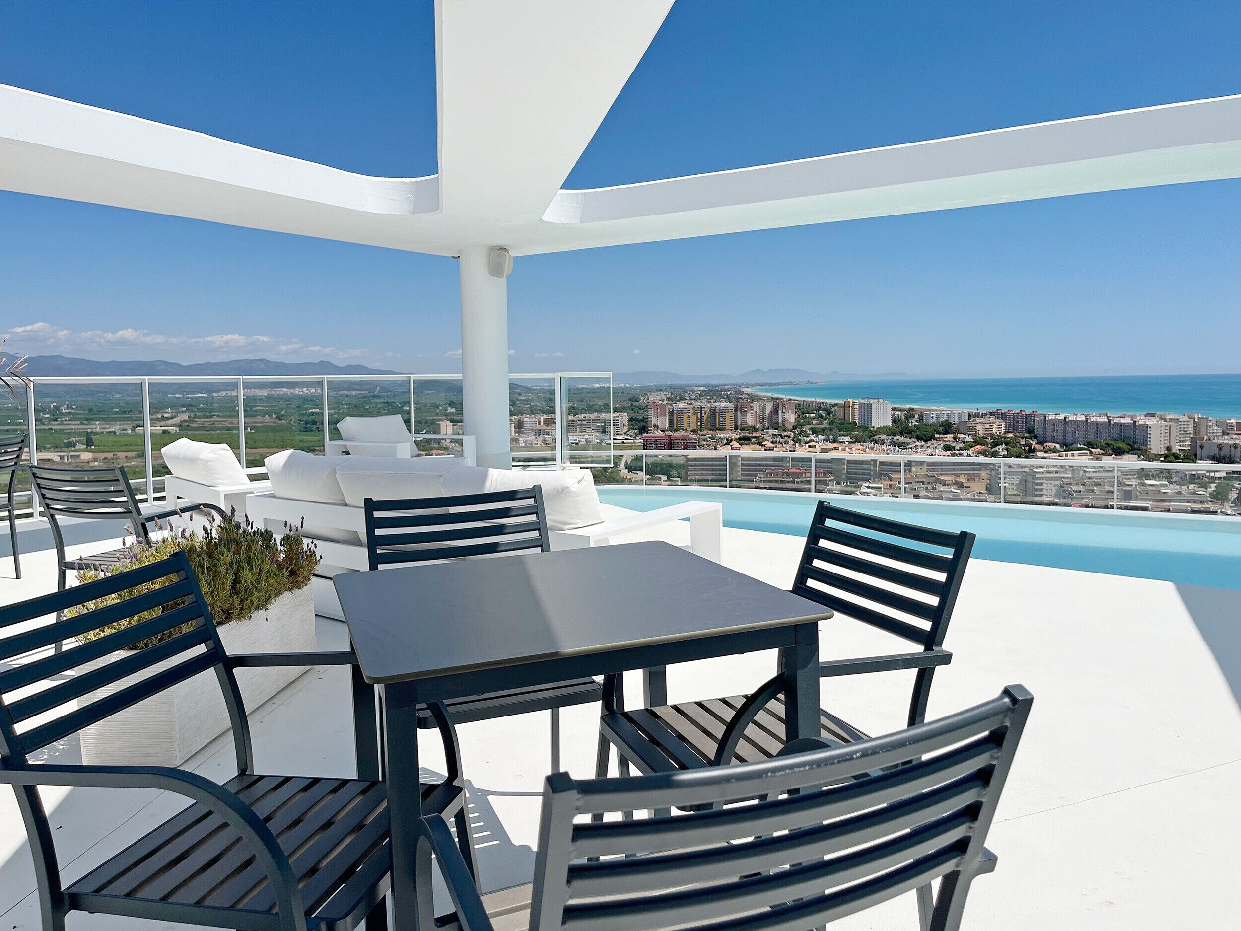 Una terraza en la azotea con piscina, mobiliario blanco y vistas a la ciudad y al océano.