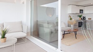 Una sala de estar moderna con un vidrio corredero que da a una cocina. Hay un sofá blanco con una mesita de noche trenzada y una planta en maceta.