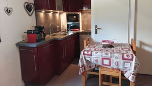 A compact kitchen with dark red cabinets, a stainless steel sink, and a microwave. A table with a patterned tablecloth is set for dining.