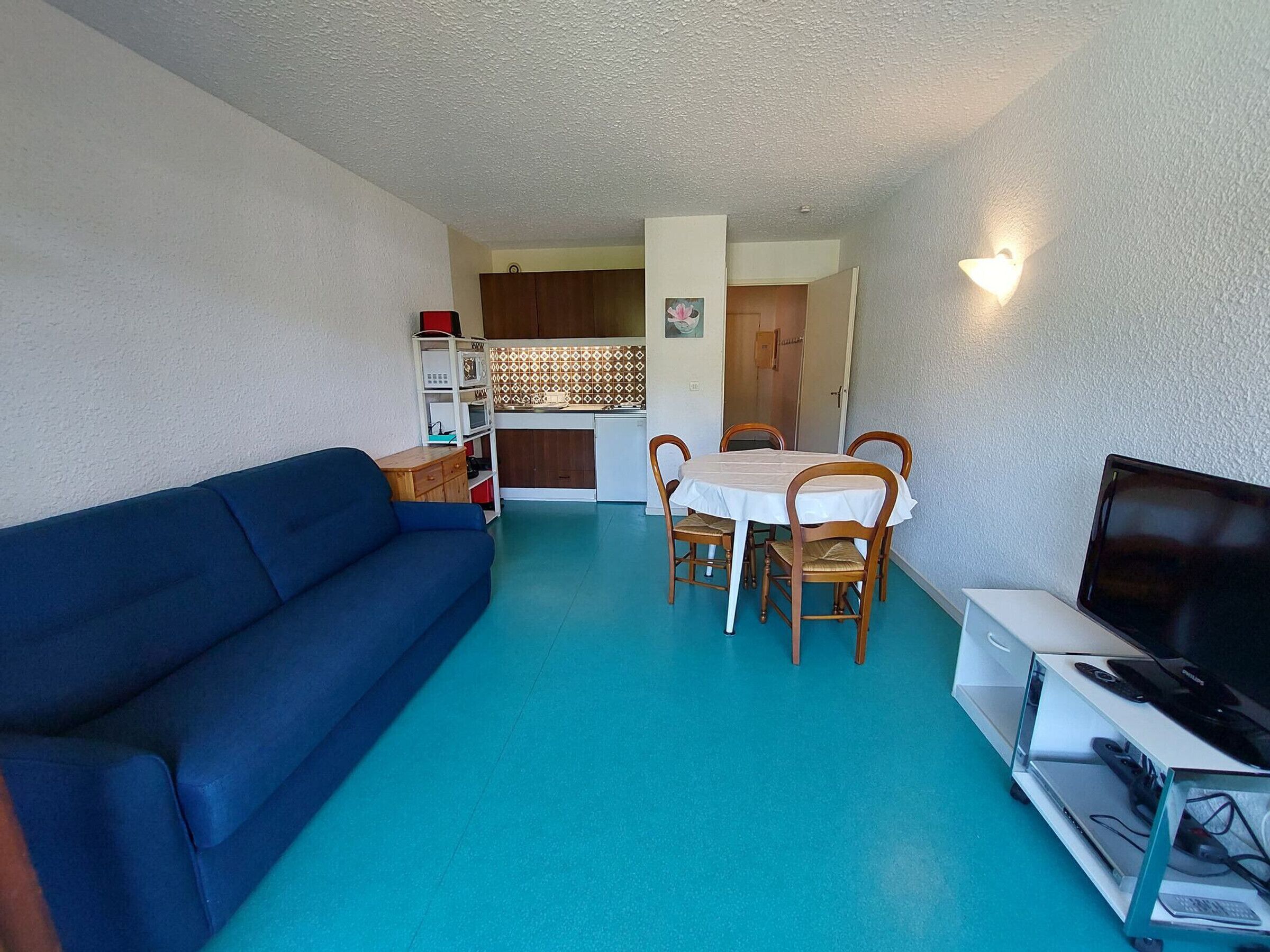 Un espacio habitable compacto con un sofá azul, una mesa de comedor con cuatro sillas, un televisor sobre un mueble bajo y una kitchenette al fondo.