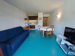 Um espaço compacto com sofá azul, mesa de jantar para quatro pessoas, televisão em suporte e uma cozinha compacta ao fundo.