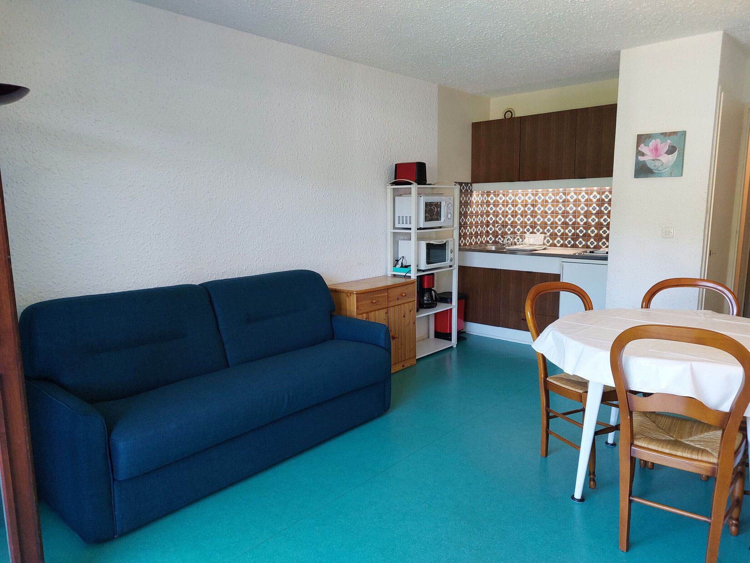 A compact living space with a blue sofa, a dining table with chairs, a kitchenette, and a small cabinet.