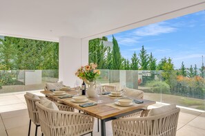 A dining area with a table set for four, surrounded by wicker chairs, with a view of greenery and trees through a glass railing. - Villa Alejandra (Benahavís)