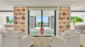 A modern living room with white sofas, a small table with a vase, and a large glass door leading to a balcony with a view of greenery. - Villa Alejandra (Benahavís)