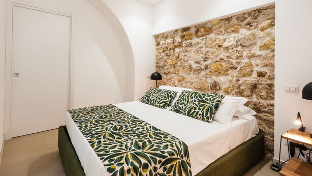 Ein Schlafzimmer mit Steinwand, einem Bett mit gemusterten Betttextilien, einer Nachttischlampe und einer Tür.