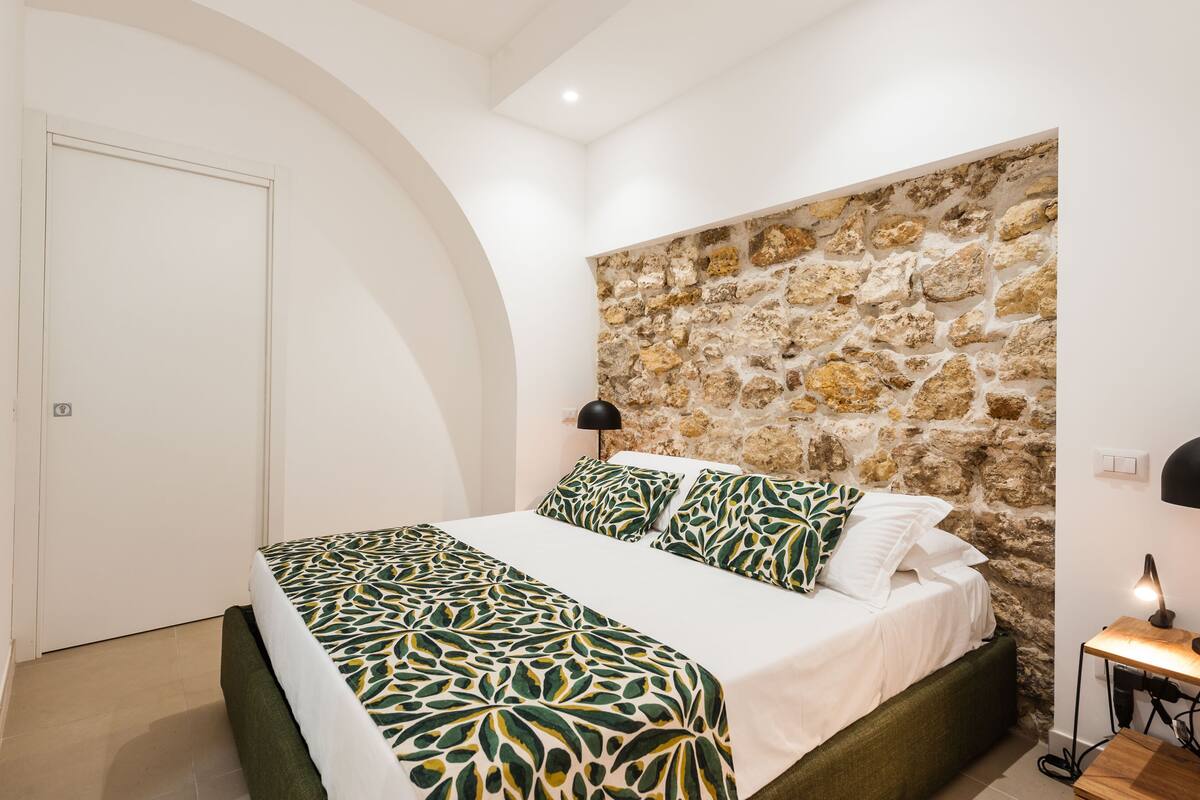 A bedroom with a stone wall, a bed with patterned bedding, a bedside lamp, and a door.