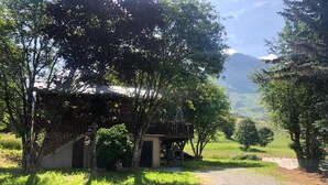Un chalet rustique doté d’une terrasse en bois, entouré d’une végétation luxuriante et accessible par un chemin de gravier.