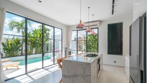 Una cocina moderna con una isla de mármol, ventanas amplias y vistas a una piscina y follaje.