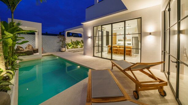 Un área de piscina moderna con una silla de descanso y un carrito de madera, rodeada por un patio bien iluminado con plantas y un espacio de comedor visible a través de puertas de vidrio.