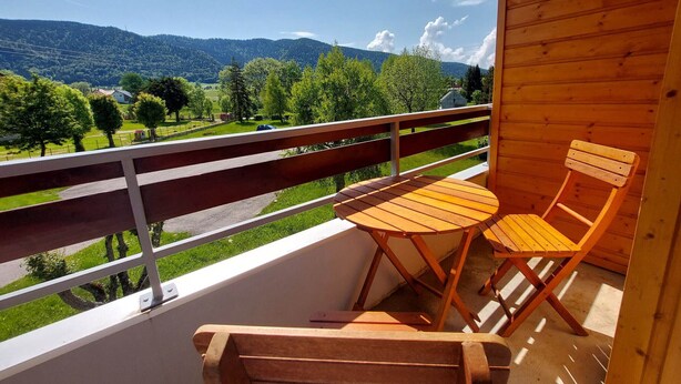 A wooden balcony with two chairs and a table, overlooking a green landscape with trees and a road.