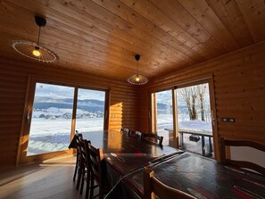 L’intérieur d’un chalet en bois, avec une grande table à manger, des chaises et deux grandes fenêtres donnant sur un paysage enneigé.