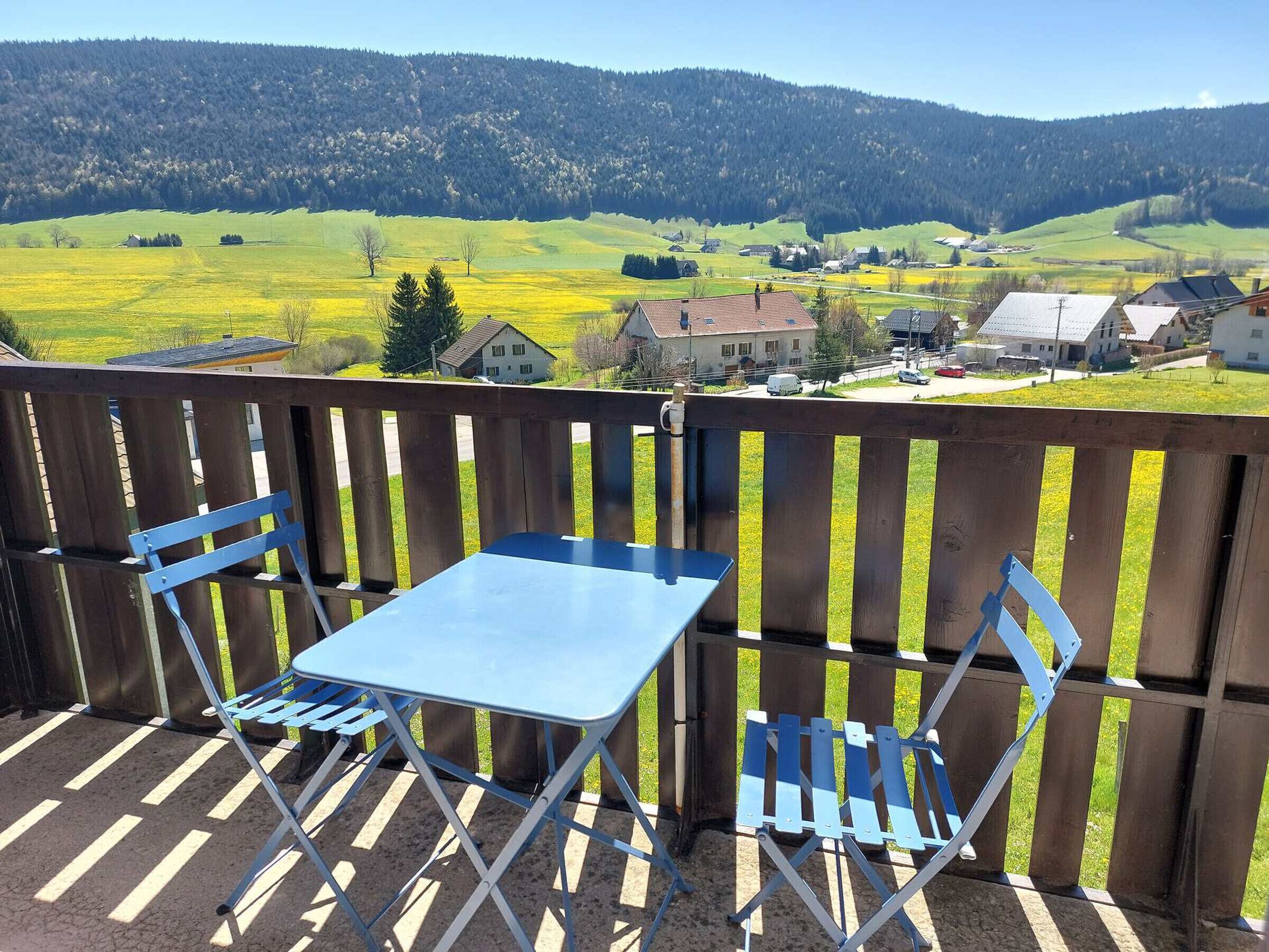 Ein Balkon mit blauem Tisch und zwei blauen Stühlen, mit Blick auf eine ländliche Landschaft mit Häusern und einem bewaldeten Hügel.