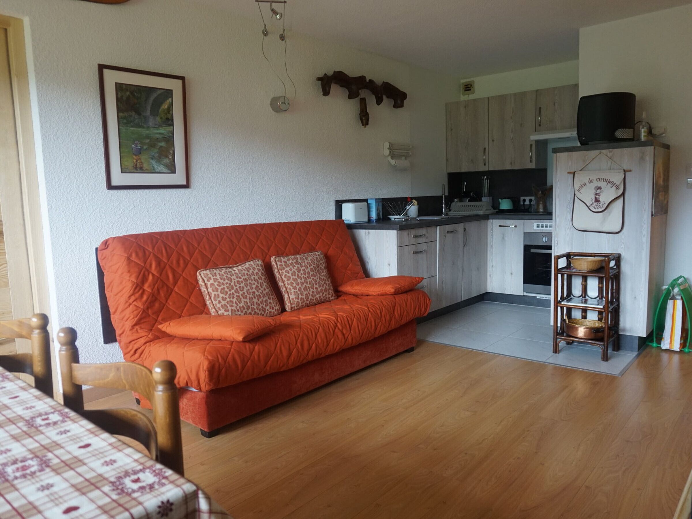 A cozy living room with a red sofa, wooden dining table, and a kitchen area with cabinets and appliances.