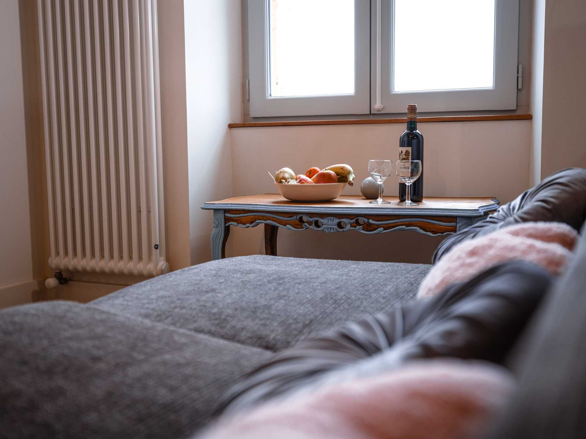 A cozy room with a bed, a table with a bowl of fruit, a bottle, and glasses.