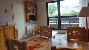 A cozy room with a wooden ceiling, a bookshelf, a desk, a chair, and a large window with a view of greenery and buildings outside. - Apartment in Méaudre - 4 pers, near center, balcony, parking (Méaudre)
