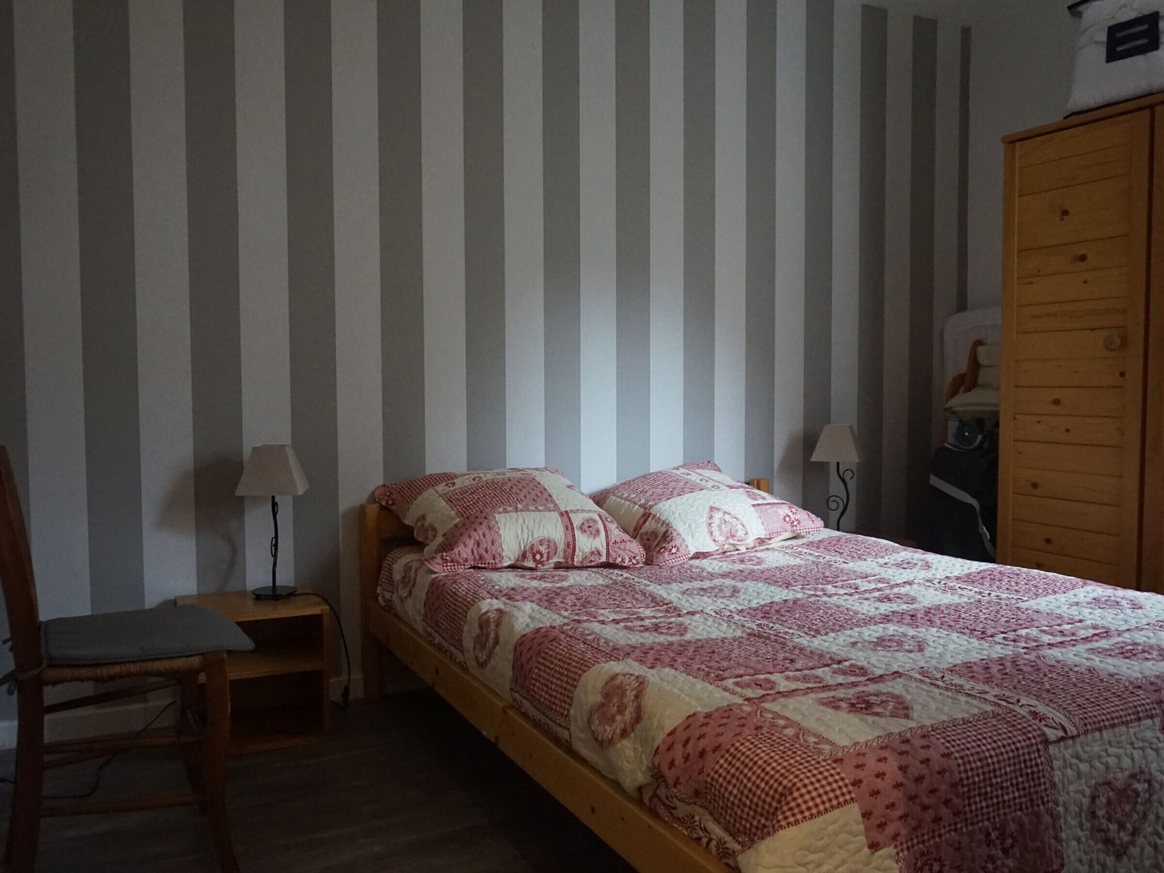 A bedroom with a wooden bed, striped wallpaper, a wooden wardrobe, and a chair.