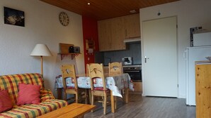 A cozy living room with a wooden sofa, a wooden coffee table, and a dining area with a table covered in a patterned tablecloth. A kitchen with wooden cabinets and a refrigerator is visible in the background. - Apartment in Méaudre - 4 pers, near center, balcony, parking (Méaudre)