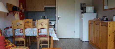 A cozy room with a bed, a wooden table, a kitchen area with cabinets and a microwave, and a door leading to another room.