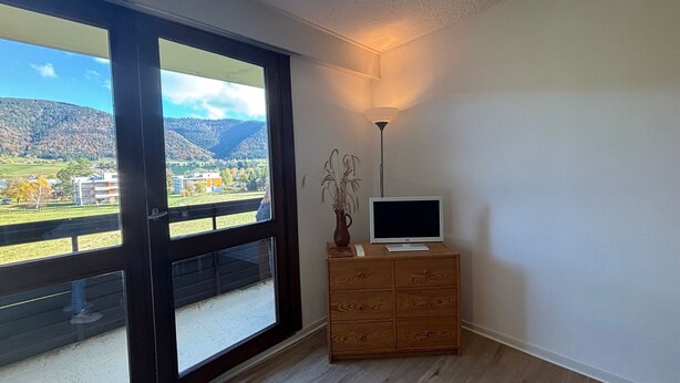 Una habitación con un gran ventanal corredero que da a un balcón con vista a la montaña. Hay un mueble de madera con un televisor de pantalla plana encima.