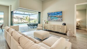 A modern living room with a large beige sectional sofa, a dining area with a glass door leading to a pool, and a large painting of a pool scene on the wall. - Villa Escape - brand new, with pool and spa, on water with gulf access (Cape Coral)