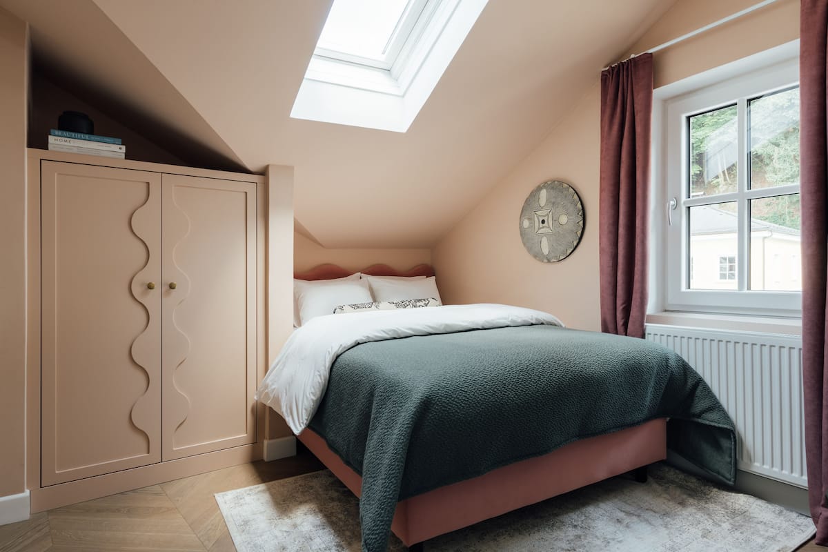 Een slaapkamer met een bed, een kledingkast, een dakraam, een raam met gordijnen en een wandklok.