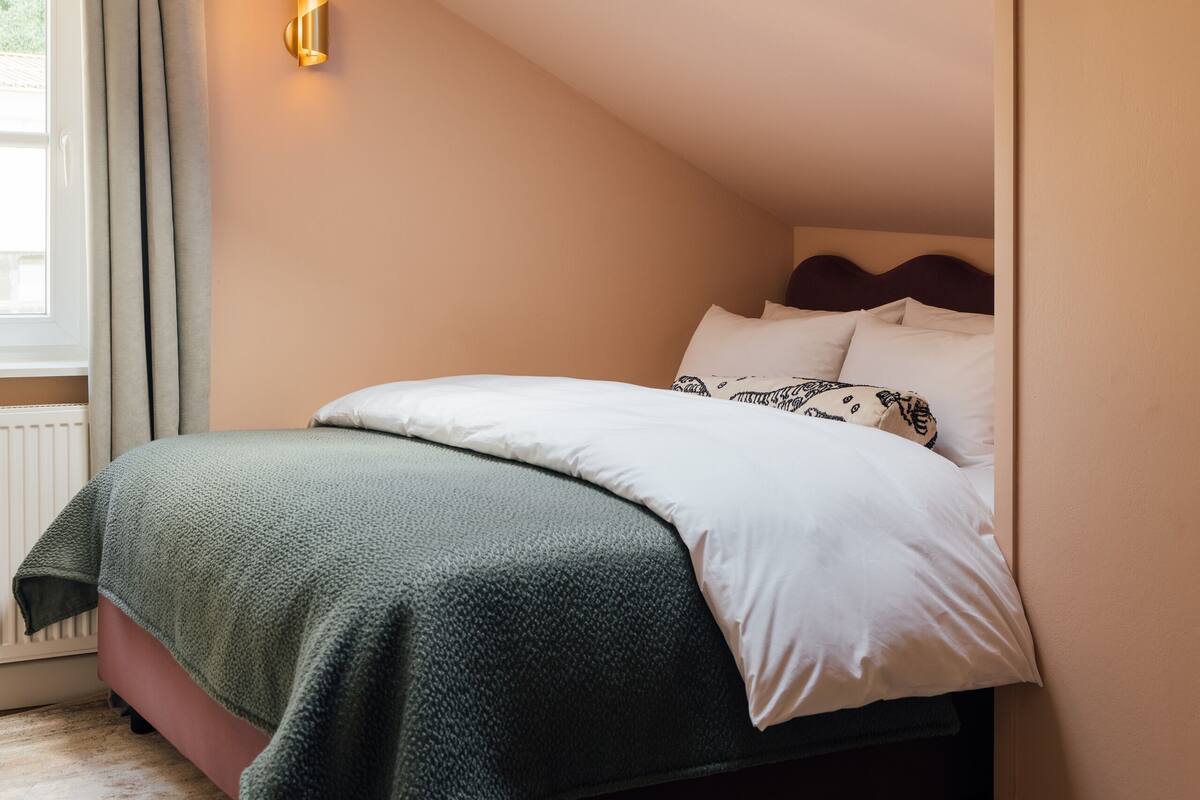 Een slaapkamer met dakraam, een bed met witte en opvallende kussens, een groene deken en een wandlamp.