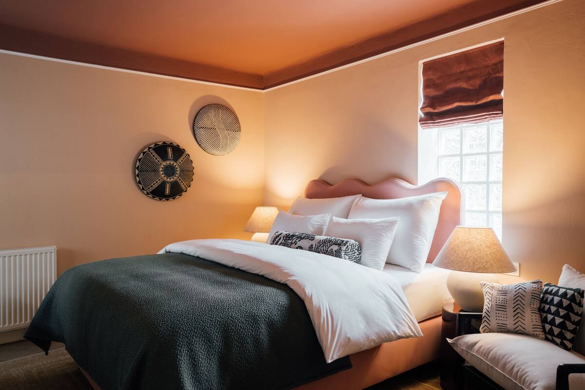 Een net opgemaakt bed met een groene deken, een bedlampje en een raam met een bruin gordijn.