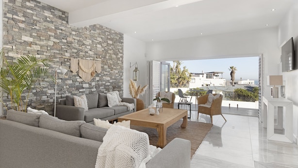 A modern living room with a stone wall, grey sofa, wooden coffee table, and a view of the city. - Endless Summer Beach House (Cape Town)