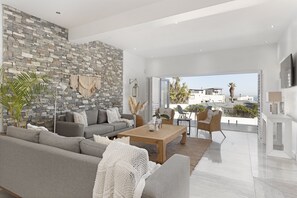 A modern living room with a stone wall, grey sofa, wooden coffee table, and a view of the city. - Endless Summer Beach House (Cape Town)