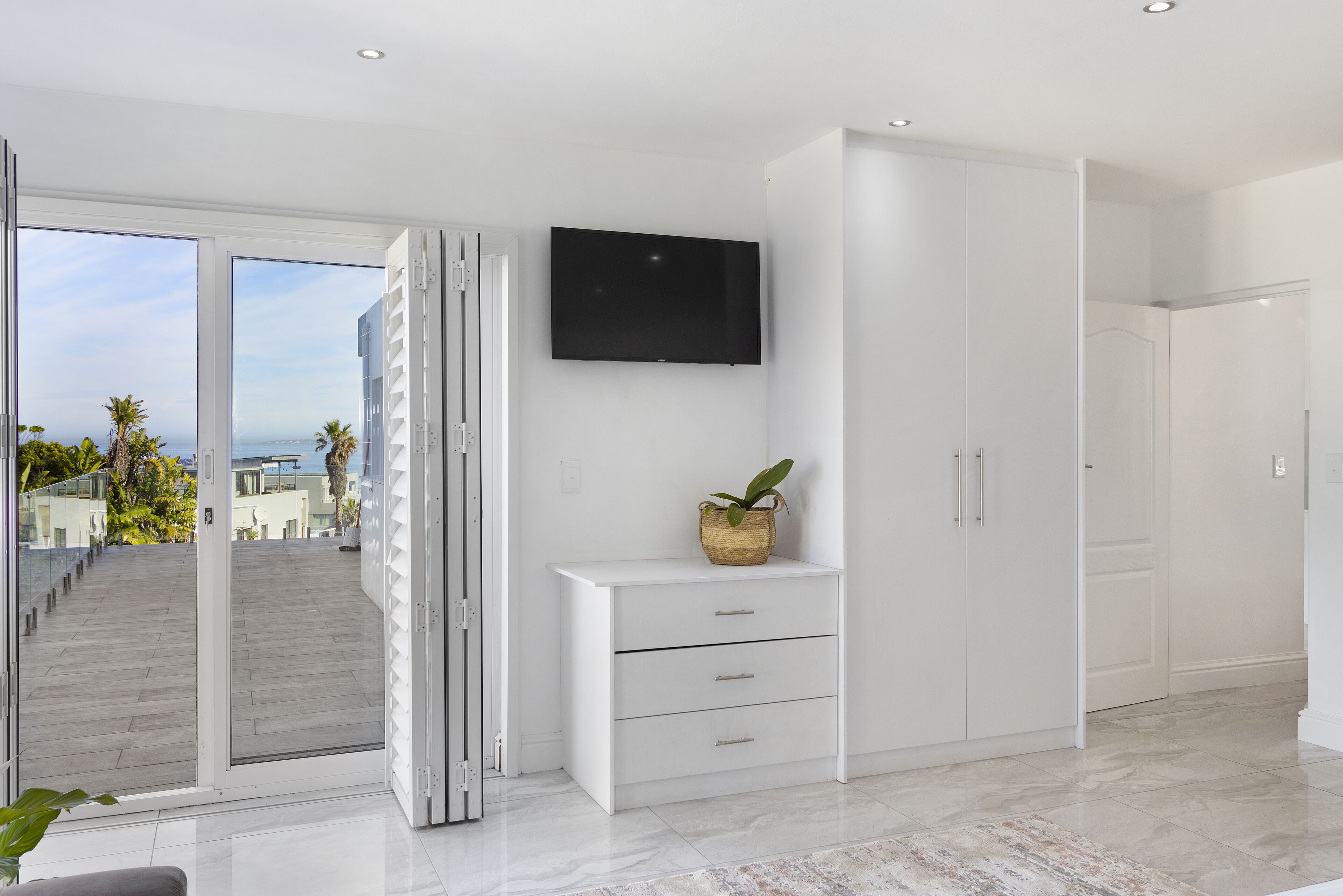 A modern hotel room with a large sliding glass door leading to a balcony, a flat-screen TV mounted on the wall, and a white console table with drawers.