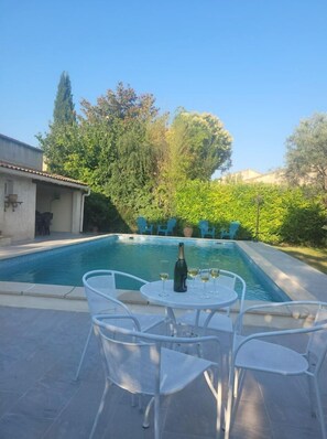 Piscina con zona de patio, mobiliario exterior y vegetación.