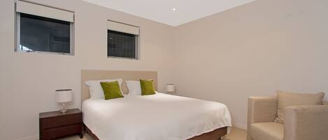 A modern hotel room with a large bed, two bedside tables, a chair, and two windows.