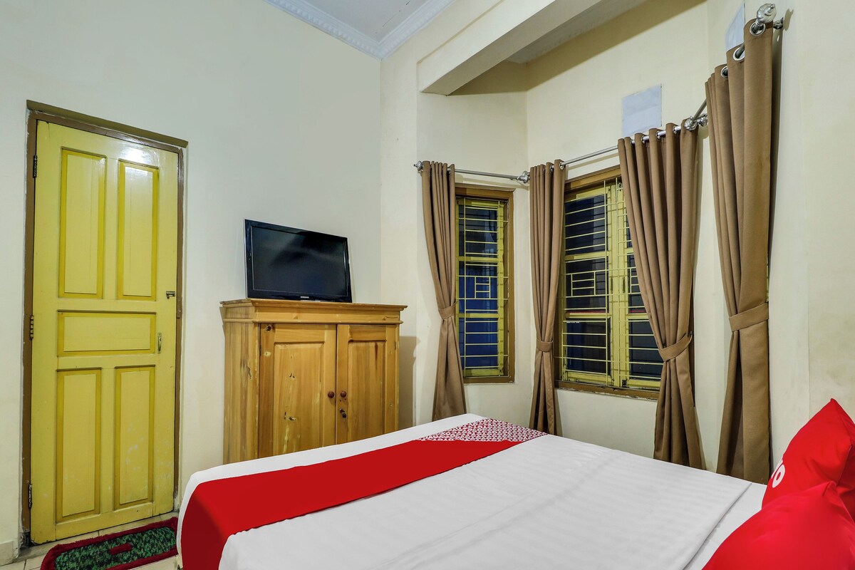 A room with a bed, a television on a wooden cabinet, and two windows with curtains.