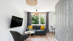 Una sala de estar moderna con un sofá gris, una pequeña mesa redonda, un sillón gris, un televisor de pantalla plana montado en la pared y un amplio ventanal con cortinas.