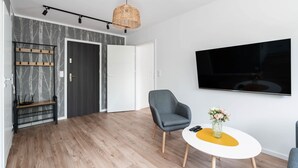 Una sala de estar moderna con un sillón gris, una mesa de centro blanca con un detalle amarillo, un televisor montado en la pared y una lámpara colgante de mimbre.