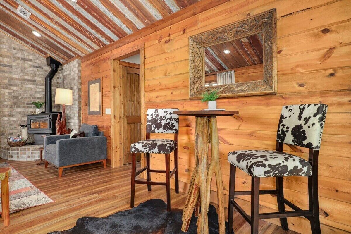 A cozy log cabin interior with a fireplace, wooden walls, a mirror, and two cowhide upholstered chairs.