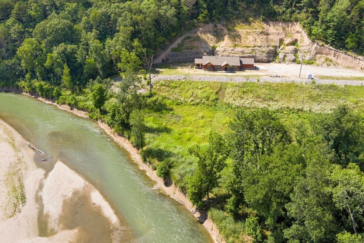 A riverside cabin with a dirt path leading to it, surrounded by dense greenery and a rocky cliff.
