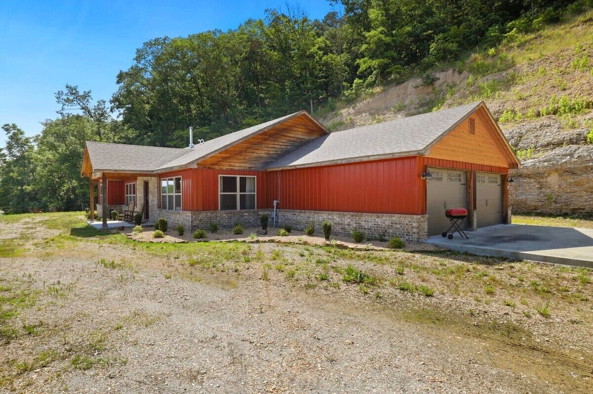A two-story building with a red exterior and a stone foundation. It features a covered porch, a garage, and a small outdoor grill area.