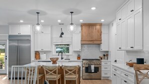 A modern kitchen with white cabinets, a stainless steel refrigerator, a wooden dining table with chairs, and pendant lights. - Three Dog Ranch - Pool, Game Room & Sleeps 18 (Driftwood)