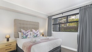Un dormitorio moderno con una cama grande, dos mesitas de noche con lámparas, una ventana con cortinas y vistas a edificios.